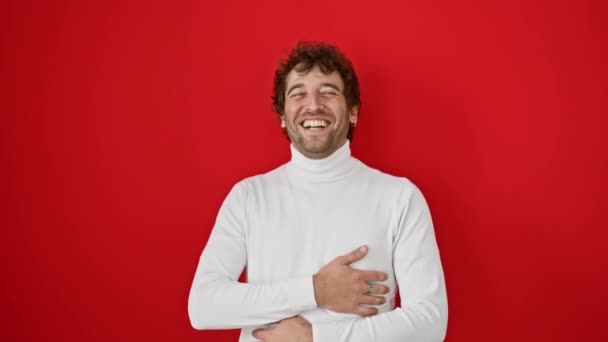 Giovane uomo ispanico allegro, in piedi in risata indossando un maglione, cintura fuori una risata dura da una battuta divertente su uno sfondo rosso isolato - Filmati, video