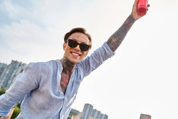 komea iloinen mies, jolla on tatuoinnit ja trendikkäät aurinkolasit, jotka poseeraavat punaisen kupin kanssa ja hymyilevät kameralle - Valokuva, kuva