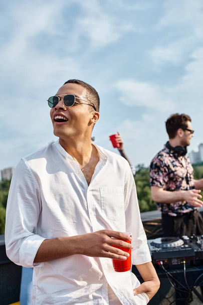 gioioso uomo africano americano con eleganti occhiali da sole in possesso di tazza rossa con bevanda alla festa sul tetto - Foto, immagini