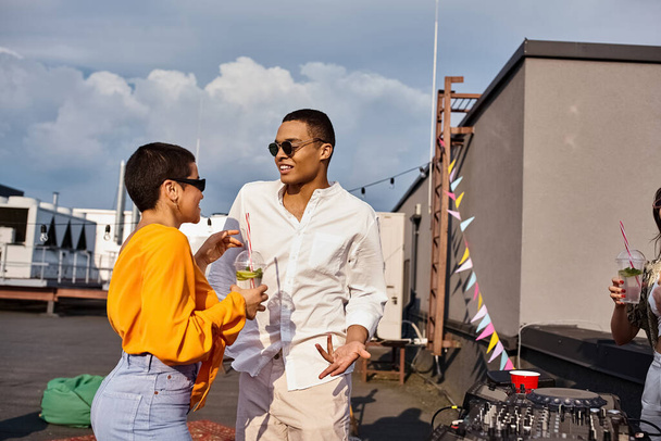 весела жінка і афроамериканський чоловік у міському одязі з сонцезахисними окулярами, що вечірки на даху - Фото, зображення