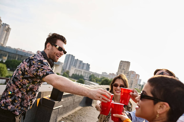 jeunes amis joyeux divers claquant leurs tasses rouges à côté de l'équipement DJ à la fête sur le toit - Photo, image