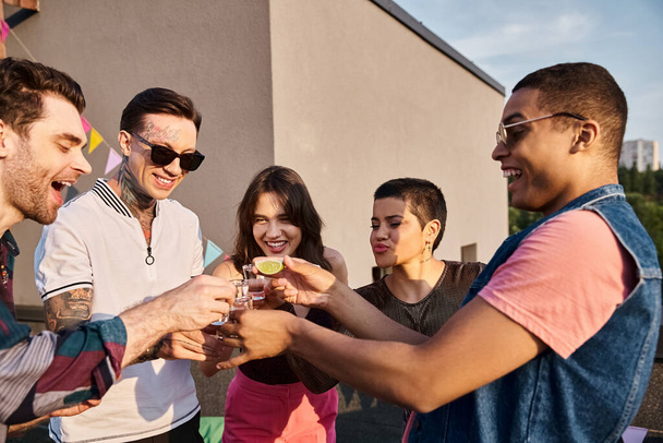 Весьма разнообразные друзья в стильных солнечных очках пьют текилу с лаймом и солью на вечеринке на крыше - Фото, изображение