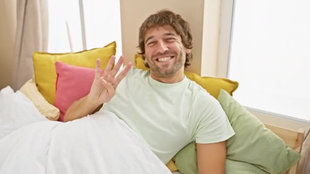 Веселый молодой кавказский парень, демонстрирующий позитивное выражение лица, уверенно сгибающий четыре пальца в спальне. в пижаме, радостно улыбаясь и указывая на идею в домашней обстановке. - Кадры, видео