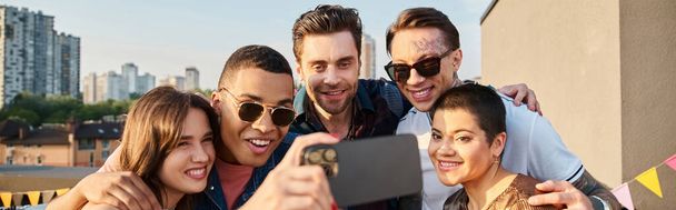 alegre interracial jóvenes amigos en casual vibrante atuendos tomando selfies en azotea partido, bandera - Foto, imagen