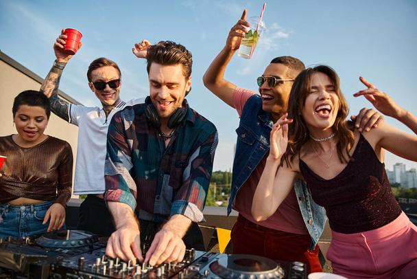 joyful good looking interracial friends in urban attires partying together with handsome DJ at party - Photo, Image