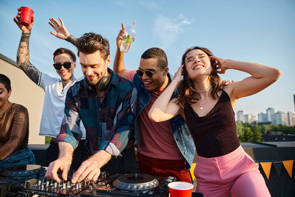 jolly multicultural people in vivid outfits partying together next to attractive DJ at rooftop party - 写真・画像