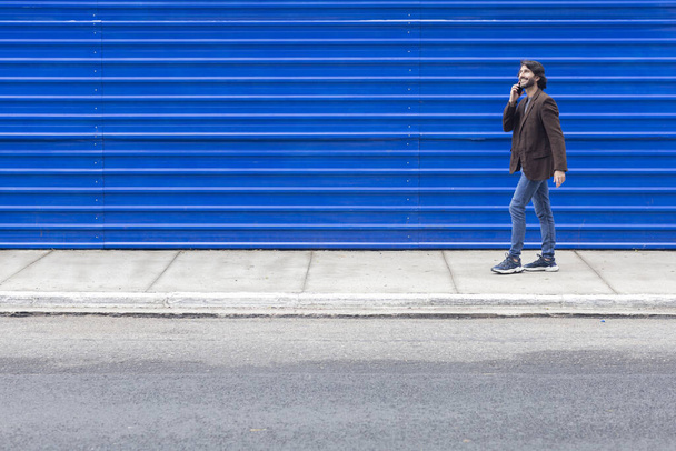 Вид на молодого чоловіка зі смартфоном, що ходить по вулиці вдень з блакитним тлом. Високоякісна фотографія. - Фото, зображення