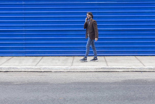 Vue d'un jeune homme avec smartphone marchant dans la rue le jour avec un fond bleu. Photo de haute qualité. - Photo, image