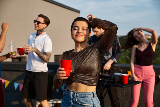 веселая молодая женщина в городском костюме держит коктейль и веселится рядом со своими разнообразными друзьями - Фото, изображение