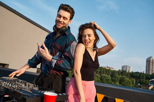 freudig attraktive Frau mit langen Haaren hat Spaß auf Dachparty neben gutaussehenden fröhlichen DJ - Foto, Bild