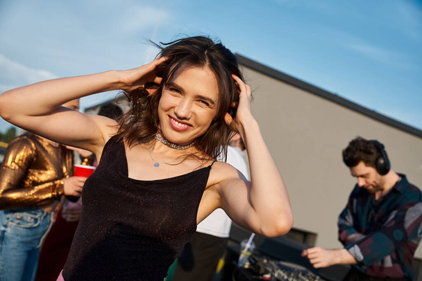 zaměření na veselou přitažlivou ženu v městském oblečení párty vedle její veselé rozmazané přátele na střeše - Fotografie, Obrázek