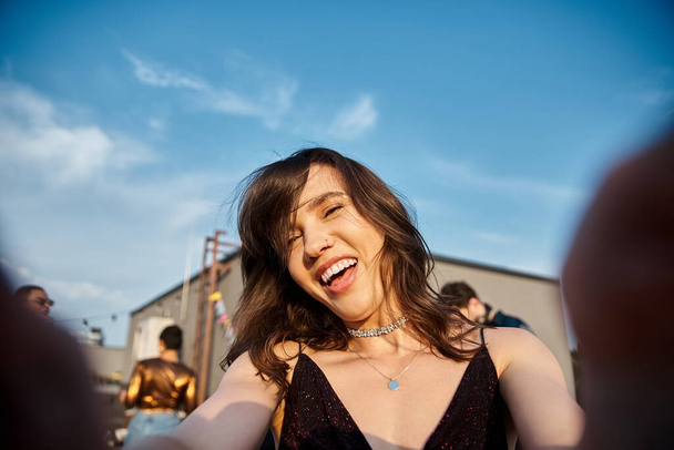 Сосредоточьтесь на веселой красивой женщине, смотрящей в камеру на вечеринке на крыше со своими разнообразными размытыми друзьями - Фото, изображение