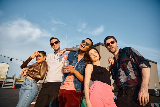 веселые привлекательные мультикультурные люди в стильных нарядах, счастливо улыбающиеся в камеру на вечеринке на крыше - Фото, изображение