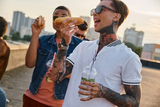 gioiosi amici multirazziali in abiti vivaci urbani godendo deliziosi hot dog sul tetto alla festa - Foto, immagini
