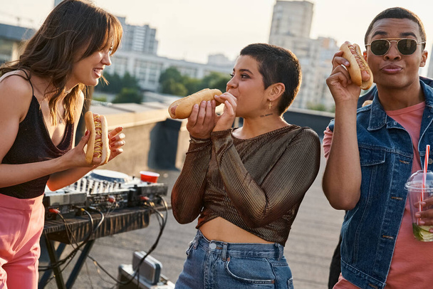 good looking joyous interracial friends in casual outfits eating tasty hot dogs at rooftop party - Photo, Image