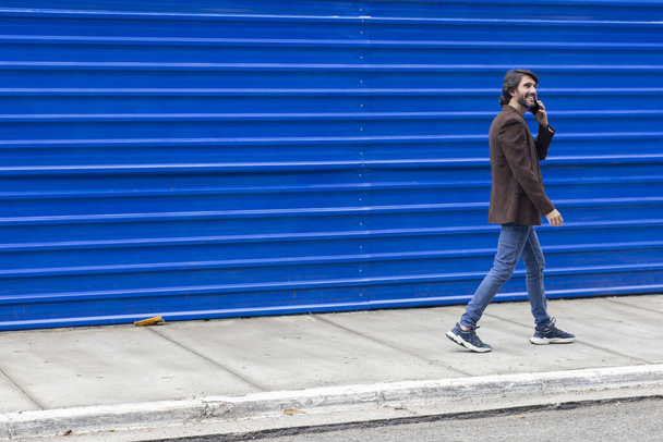 Вид молодого человека со смартфоном, идущего по улице днем на синем фоне. Высокое качество фото. - Фото, изображение