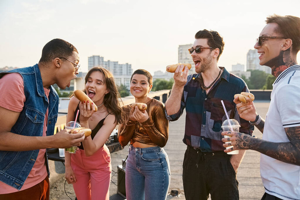 gut aussehende fröhliche diverse Freunde in lässigen Outfits essen leckere Hot Dogs auf der Dachparty - Foto, Bild