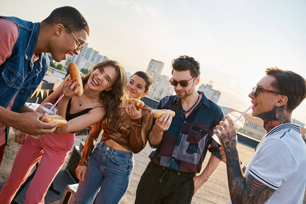cheerful multicultural people in vivid outfits enjoying delicious hot dogs on rooftop at party - Photo, Image