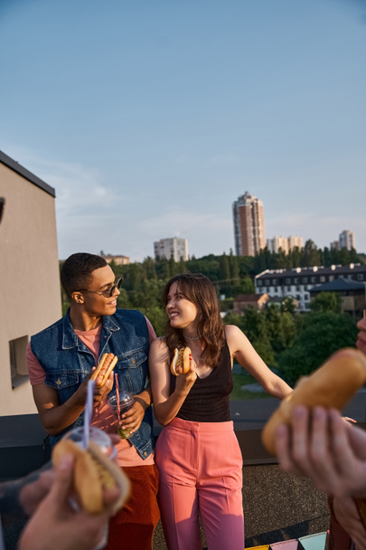 веселі багаторасові друзі у яскравих нарядах насолоджуються смачними хот-догами на даху на вечірці - Фото, зображення