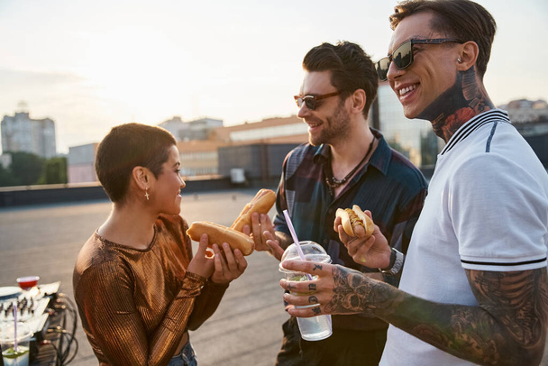 radosny dobrze wyglądający przyjaciele z okularami przeciwsłonecznymi w miejskich strojach cieszący hot dogi na dachu party - Zdjęcie, obraz