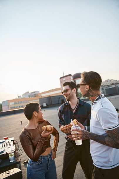 joyful good looking people with sunglasses in vivid outfits enjoying hot dogs at rooftop party - Photo, Image