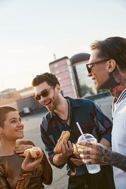 wesoły atrakcyjne przyjaciele z okularami przeciwsłonecznymi w miejskich strojach korzystających hot dogi na dachu party - Zdjęcie, obraz