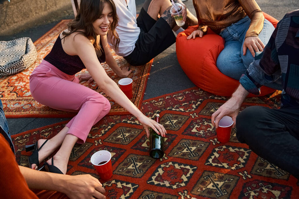 fröhliche junge multikulturelle Freunde in lebendigen Gewändern spielen Flaschendrehen bei Dachparty - Foto, Bild