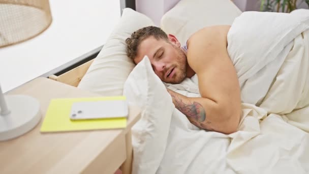 Baard man wakker gedesoriënteerd in een moderne slaapkamer, reikend naar zijn smartphone op het nachtkastje. - Video