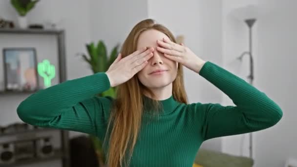 Cheerful young blonde woman sparkles at home, confidently wearing a sweater and covering her eyes with her hands for some playful blind fun. her radiant smile lights up the room! - Footage, Video