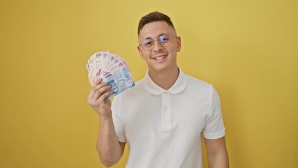 Веселый молодой латиноамериканец, уверенно держащий мексиканские банкноты песо, жестикулирующий отличным знаком "ок" пальцем, излучающий успех на изолированном желтом фоне. - Кадры, видео