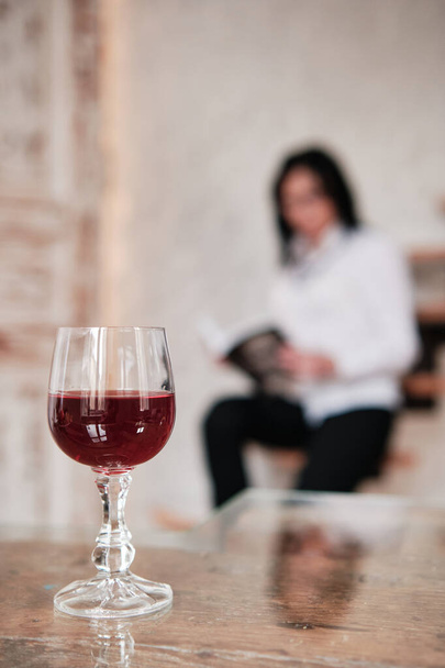 Un bicchiere di vino rosso prende il centro della scena, con una donna che legge un libro sullo sfondo, creando un senso di relax e intrigo. Focus selettivo - Foto, immagini