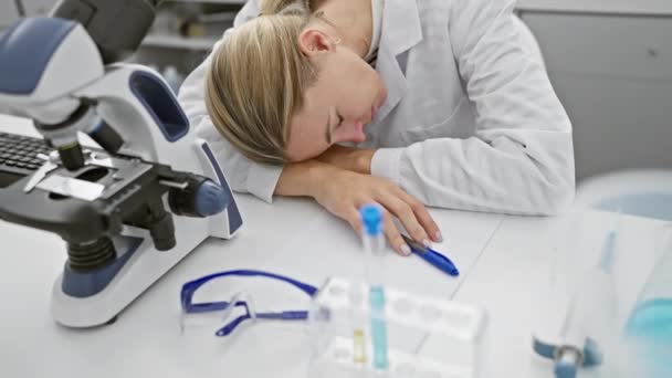 Κουρασμένη νεαρή καυκάσια γυναίκα επιστήμονας σε lab coat εργάζονται με ένα μικροσκόπιο σε ένα εργαστηριακό περιβάλλον. - Πλάνα, βίντεο