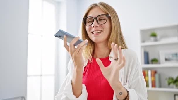 Een jonge vrouw in een kantoor spreekt in een smartphone, gebaren terwijl ze een bril draagt, een witte jas en een rode top. - Video