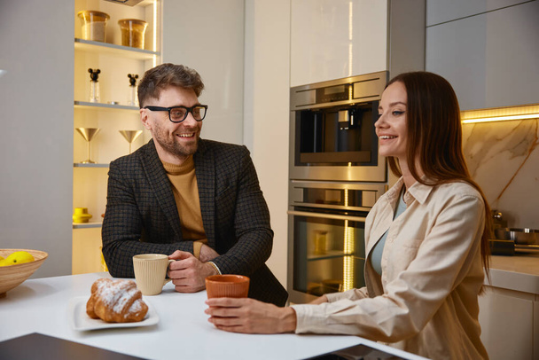 Happy romantic couple drinking coffee talking sharing day news at modern home kitchen with luxury interior. Stylish handsome man and attractive woman spending time together - Photo, Image
