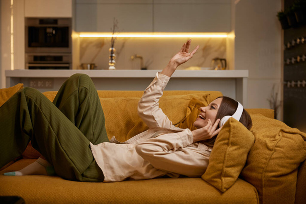 Щаслива розслаблена молода жінка слухає музику в навушниках вдома. Тисячолітня жінка лежить на зручному дивані - Фото, зображення