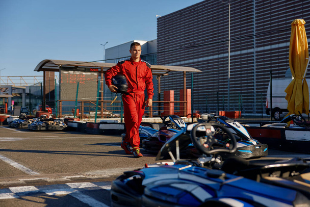 Well-equipped go-kart driver wearing overalls and holding helmet walking to car before race at starting line on motor racing track. Automotive sports activity, carting championship - Photo, Image