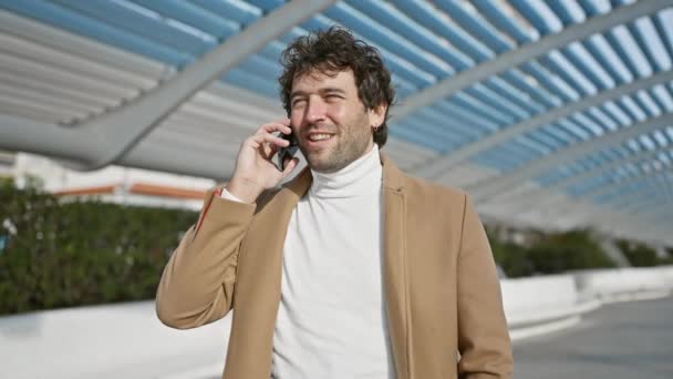 Un hombre hispano con estilo habla y reacciona a una conversación en su teléfono inteligente fuera bajo la arquitectura moderna. - Metraje, vídeo