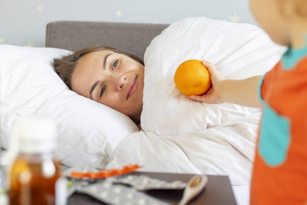 Un bambino premuroso porge un'arancia alla madre malata sdraiata a letto, con farmaci visibili sul comodino.. - Foto, immagini