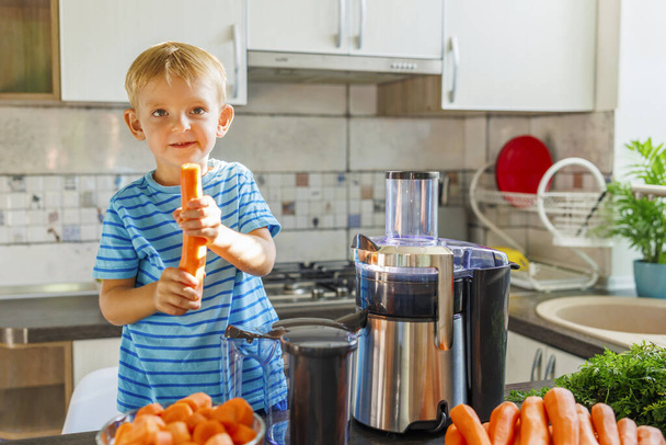 Uśmiechnięty chłopiec przygotowuje sok marchewkowy używając sokowirówki w słonecznej kuchni. - Zdjęcie, obraz