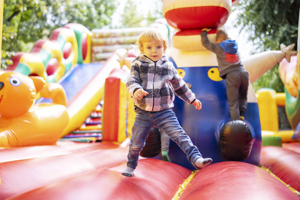 Criança nova que joga em um castelo bouncy inflável vibrante em um campo de jogos exterior cercado pela vegetação. - Foto, Imagem