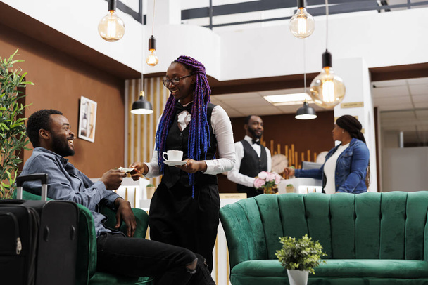 Elégedett afro-amerikai turista, aki pénzt ad a pincérnőnek, miközben a hotel halljában ül csomagokkal. Utazó kávét vásárol, miközben a vendég bejelentkezésére vár - Fotó, kép