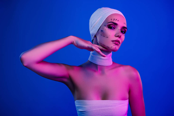 Foto di attraente ragazza toccare volto mento con segni di correzione per la chirurgia sguardo su sfondo blu al neon. - Foto, immagini