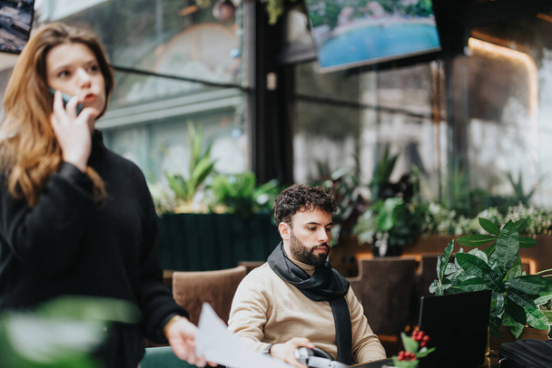 Skupiony mężczyzna z laptopem i kobietą rozmawiający przez telefon w bujnej kawiarni, ucieleśniający nowoczesną elastyczność pracy i atmosferę pracy. - Zdjęcie, obraz