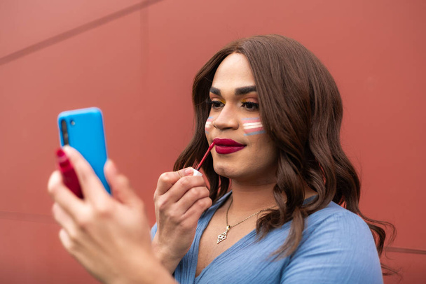 Mujer trans joven que se pone maquillaje y lápiz labial usando el teléfono celular como espejo. Concepto de estilo de vida LGTB. Foto de alta calidad - Foto, imagen