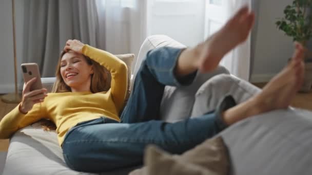 Onnellinen nainen soittaa kännykkään kotona. Rento positiivinen nainen tehdyn sohva tehdä selfie matkapuhelimeen. Hymyilevä tyttö tilalla älypuhelin nauttia laiska viikonloppuna valossa huoneisto  - Materiaali, video