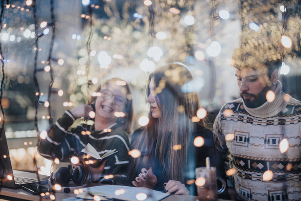 Un grupo de adultos jóvenes se dedican a un esfuerzo de colaboración alrededor de un ordenador portátil en un café calurosamente iluminado adornado con luces de hadas, exudando un ambiente acogedor. - Foto, Imagen
