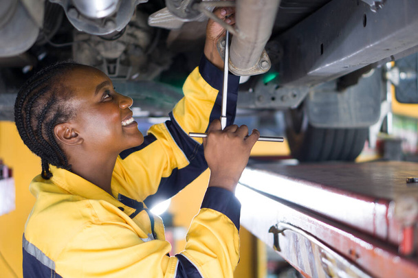 Eine Frau in Uniform arbeitet unter einem Auto, das auf einem hydraulischen Hubgestell in einer Kfz-Werkstatt angehoben wird. Der Mechaniker hält ein Werkzeug fest oder löst ein Bauteil unter dem Fahrzeug. - Foto, Bild
