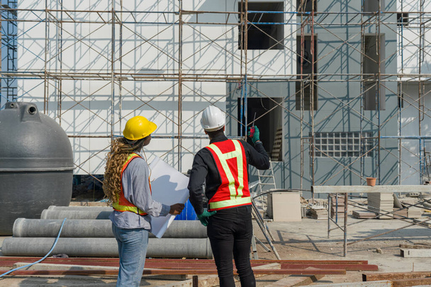 Двое строителей стоят вместе перед большой строительной площадкой со шлемом и защитным снаряжением. Дневная проверка безопасности труда. Рабочая среда на площадке жилищных проектов. - Фото, изображение