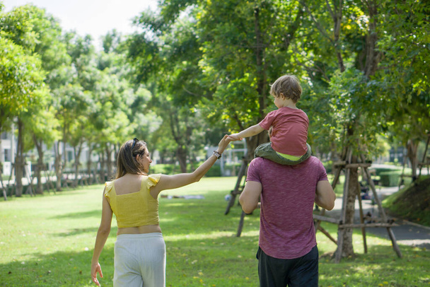 Uma mulher, seu marido e seu filho caminham juntos, desfrutando da natureza em um parque ensolarado com árvores e um caminho a pé. Vista por trás - Foto, Imagem