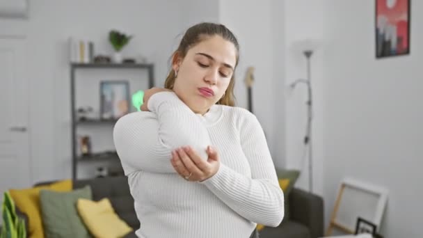 Jeune femme hispanique en pull blanc éprouvant une douleur au coude à l'intérieur d'une maison confortable - Séquence, vidéo